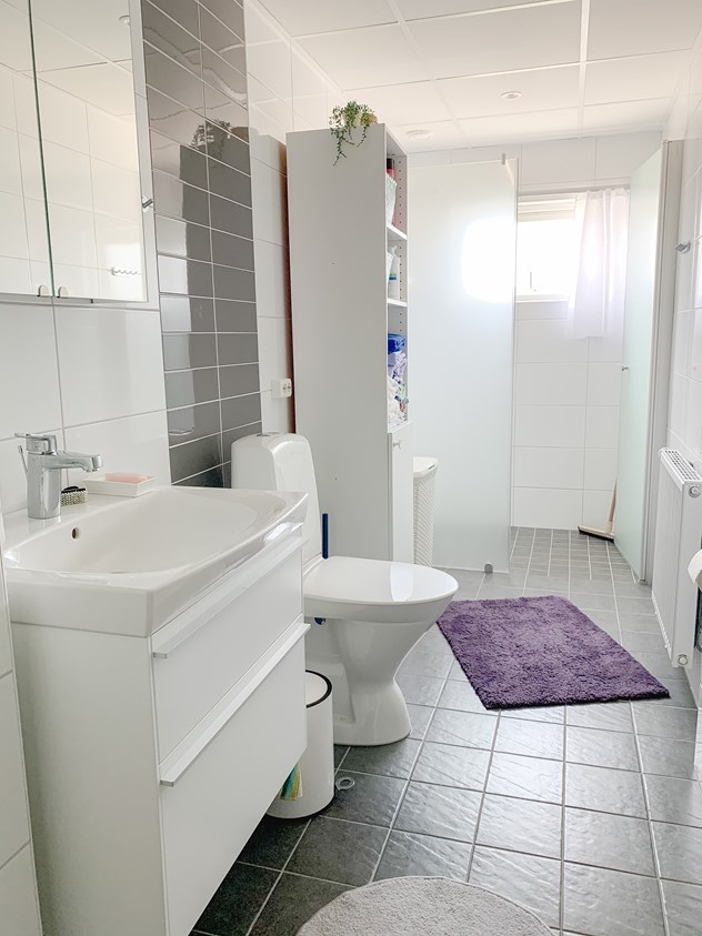 Nyrenoverat badrum med grått klinkel och vitt kakel. Ny vit WCstol och vitt tvättställ. På golvet ligget en lila matta