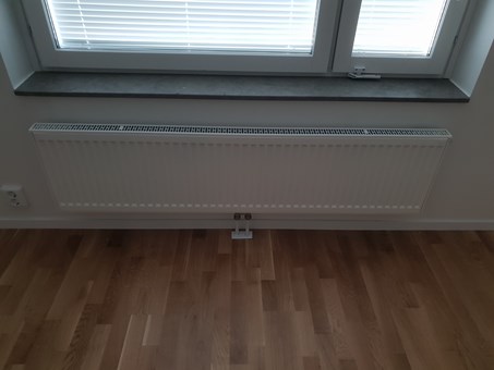 Vit radiator under ett vitt fönster. Fönstret har persienner. Golvet är nylagt av parkett.